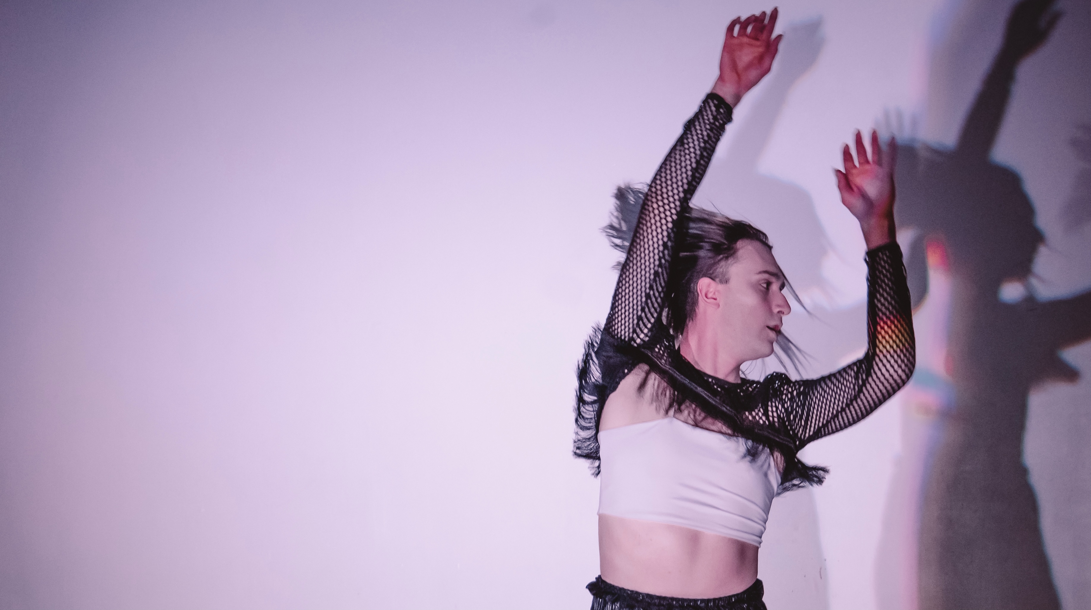 A dancer seen from the waist up against a solid background tosses their arms freely up. They wear a white bandeau and black mesh sleeves with fringe and their bleached blonde hair flying in motion.