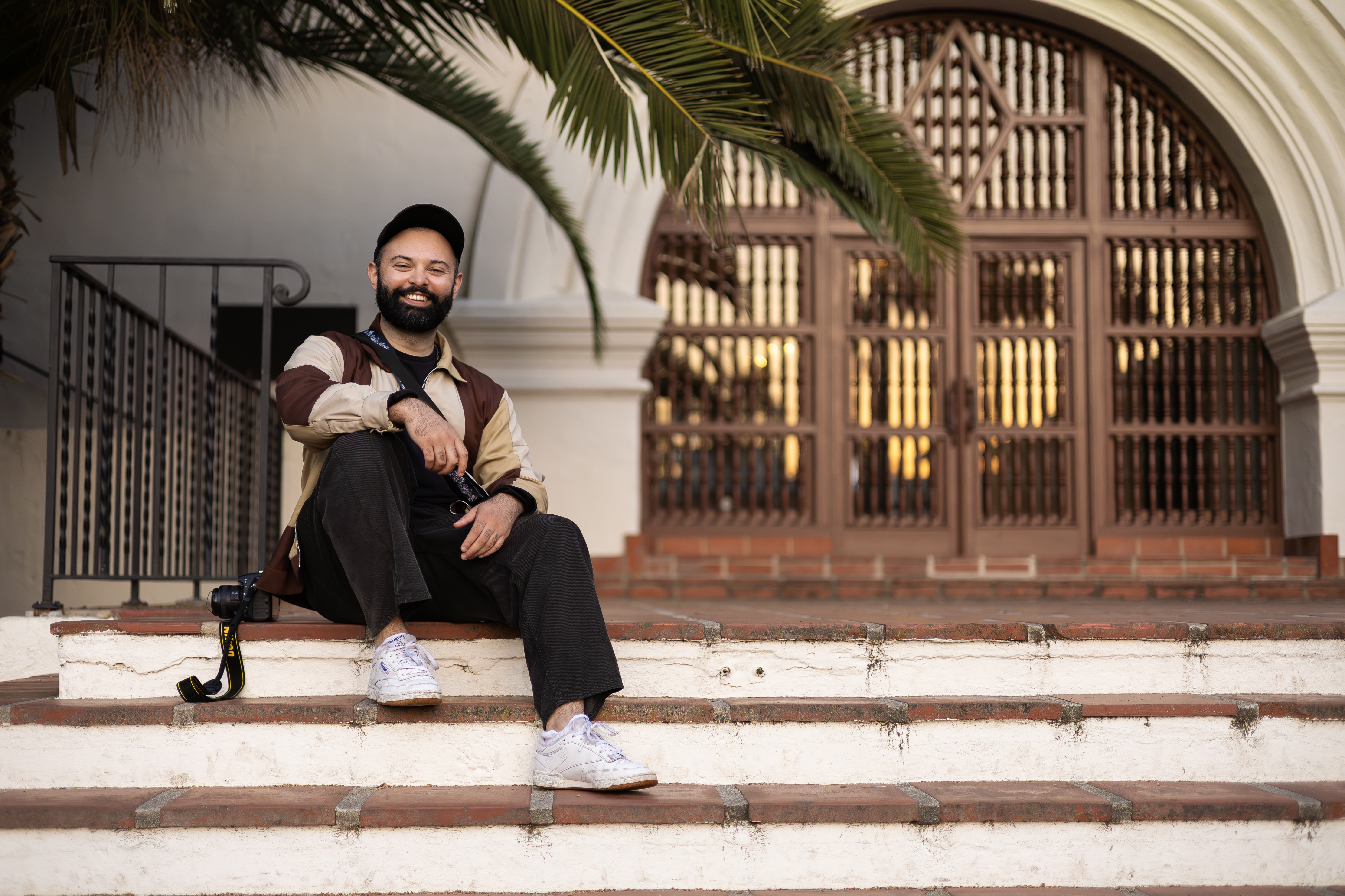 A young, light mocha-skinned man sits on a set of Spanish-style stairs wearing a ball cap and oversized canvas coat