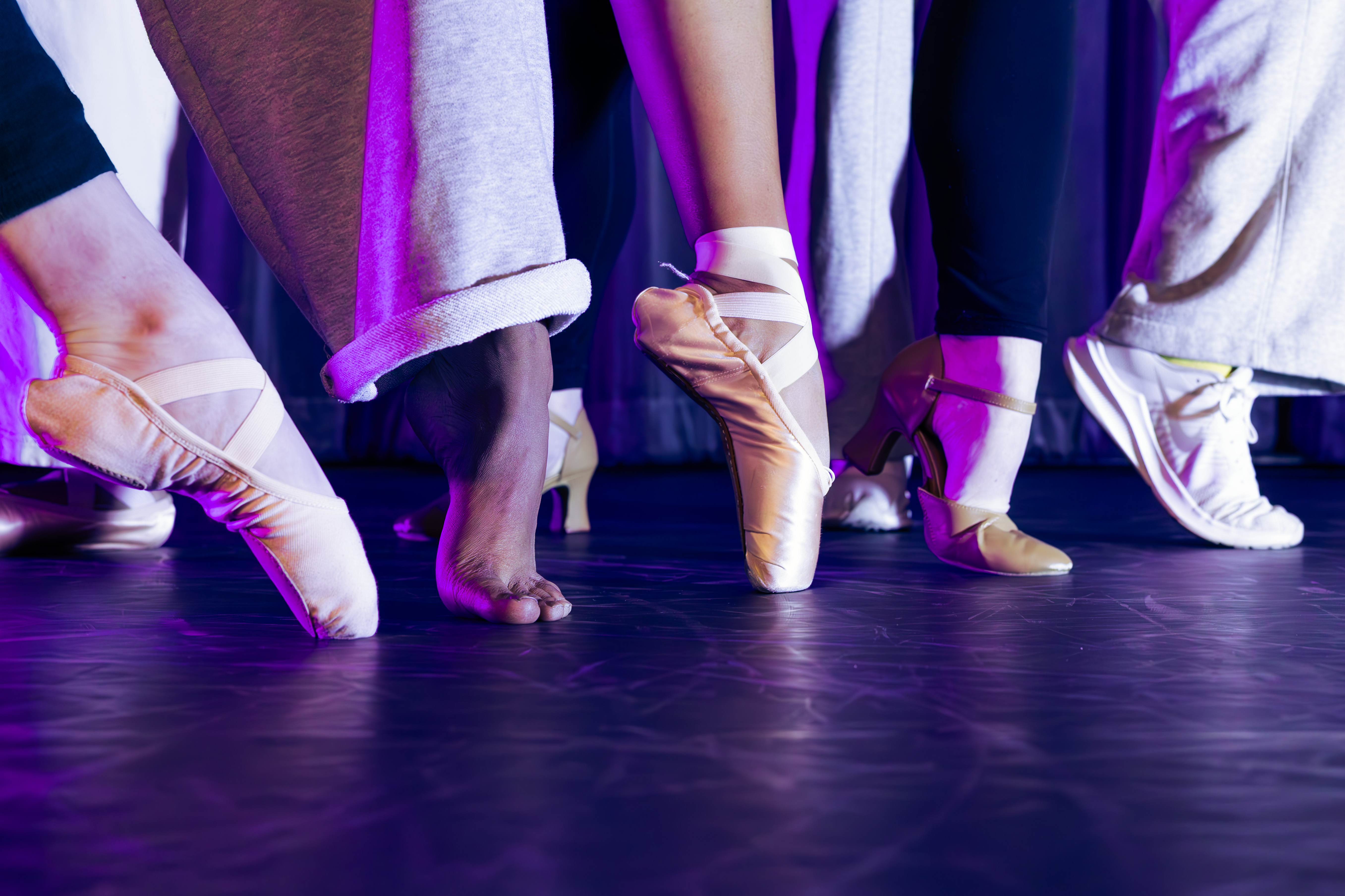 Five dancers feet are seen in profile wearing different types of dance shoes