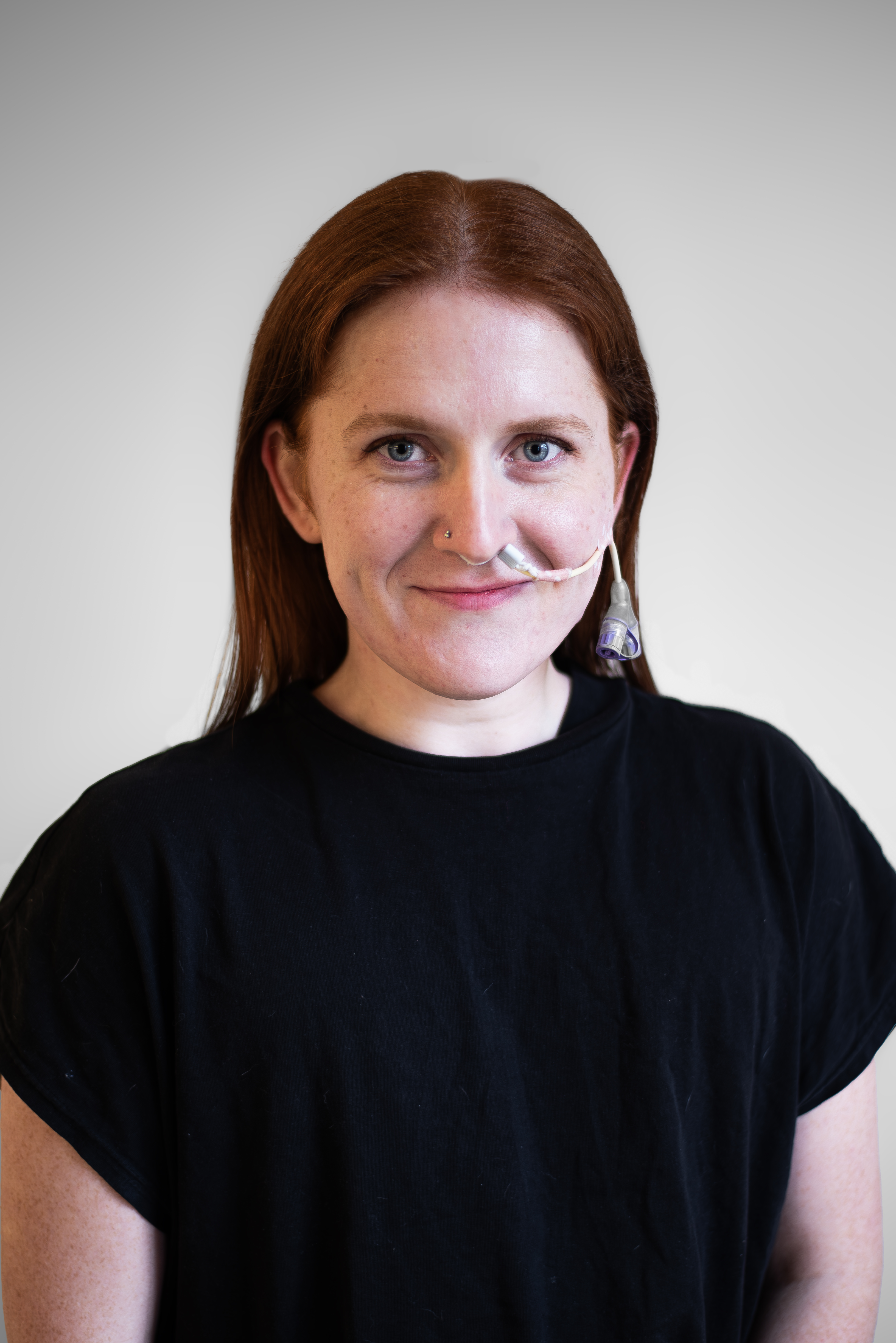 a young person with fair skin and shoulder length red hair smiles at the camera sitting against a blank wall