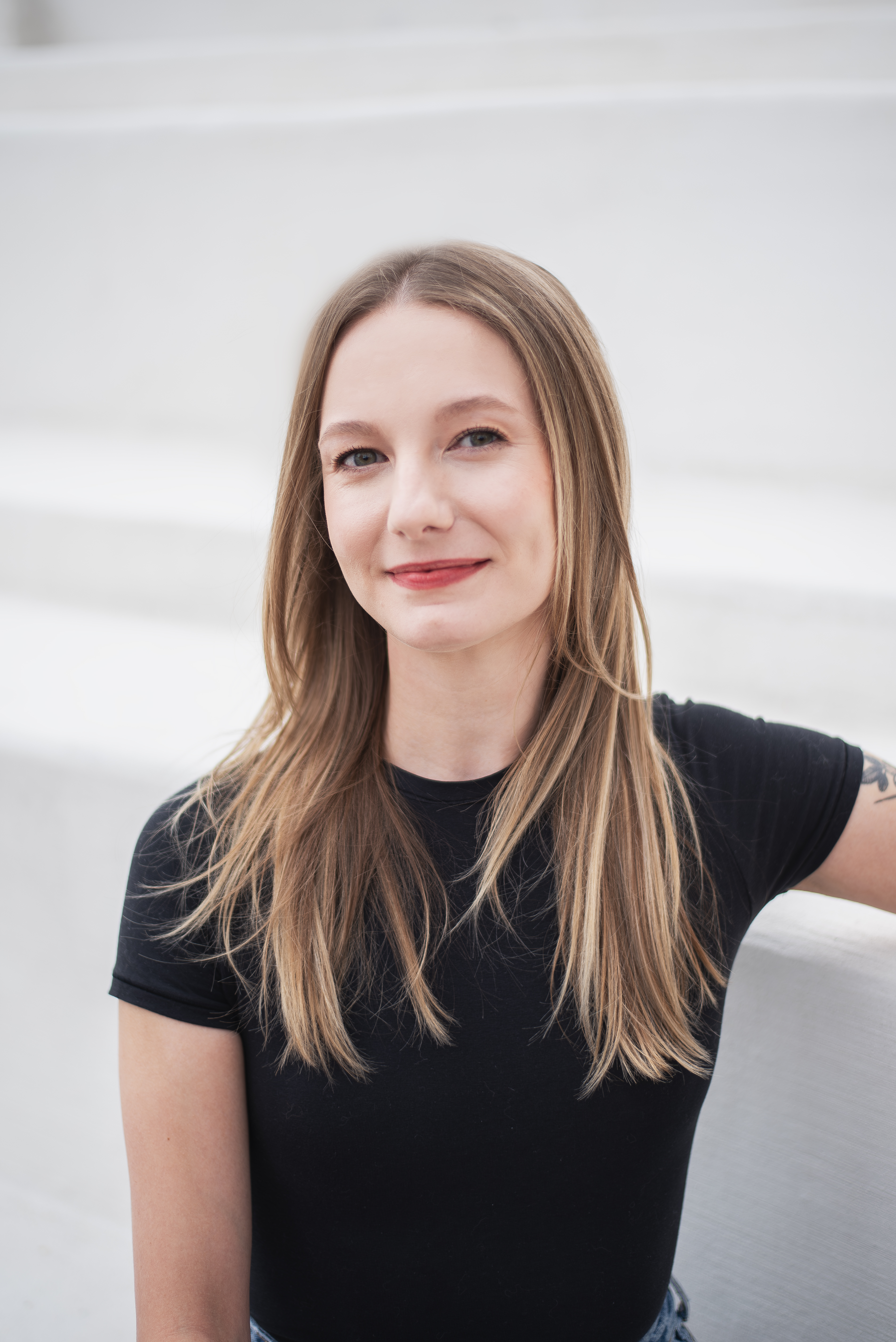 a young blonde woman is seen in close-up with fair skin and a deep red lip half-smiles at the camera as she sits in a large, white amphitheater