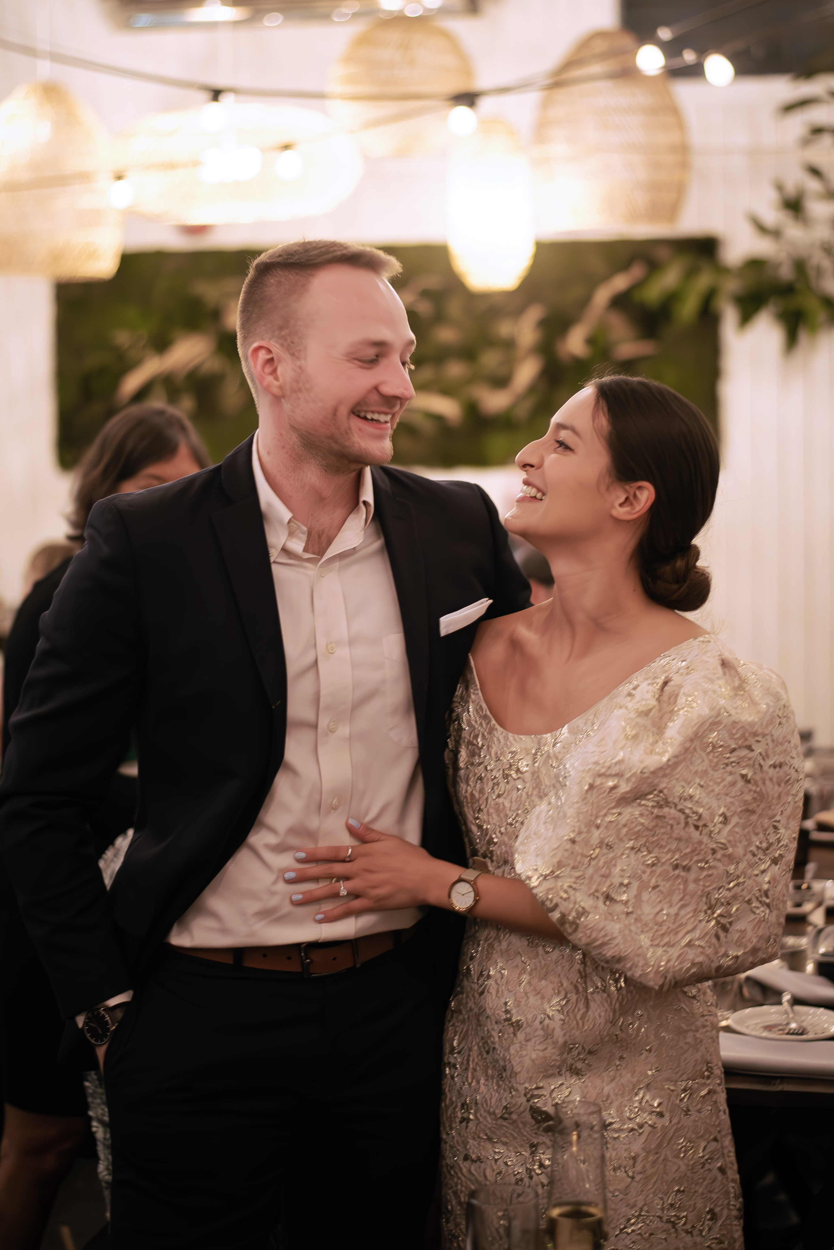 a young man with cropped blonde hair and fair skin in a suit stands next to a young woman with warm olive skin, her brown hair in a low bun pulled back from her white puffy-sleeved dress.