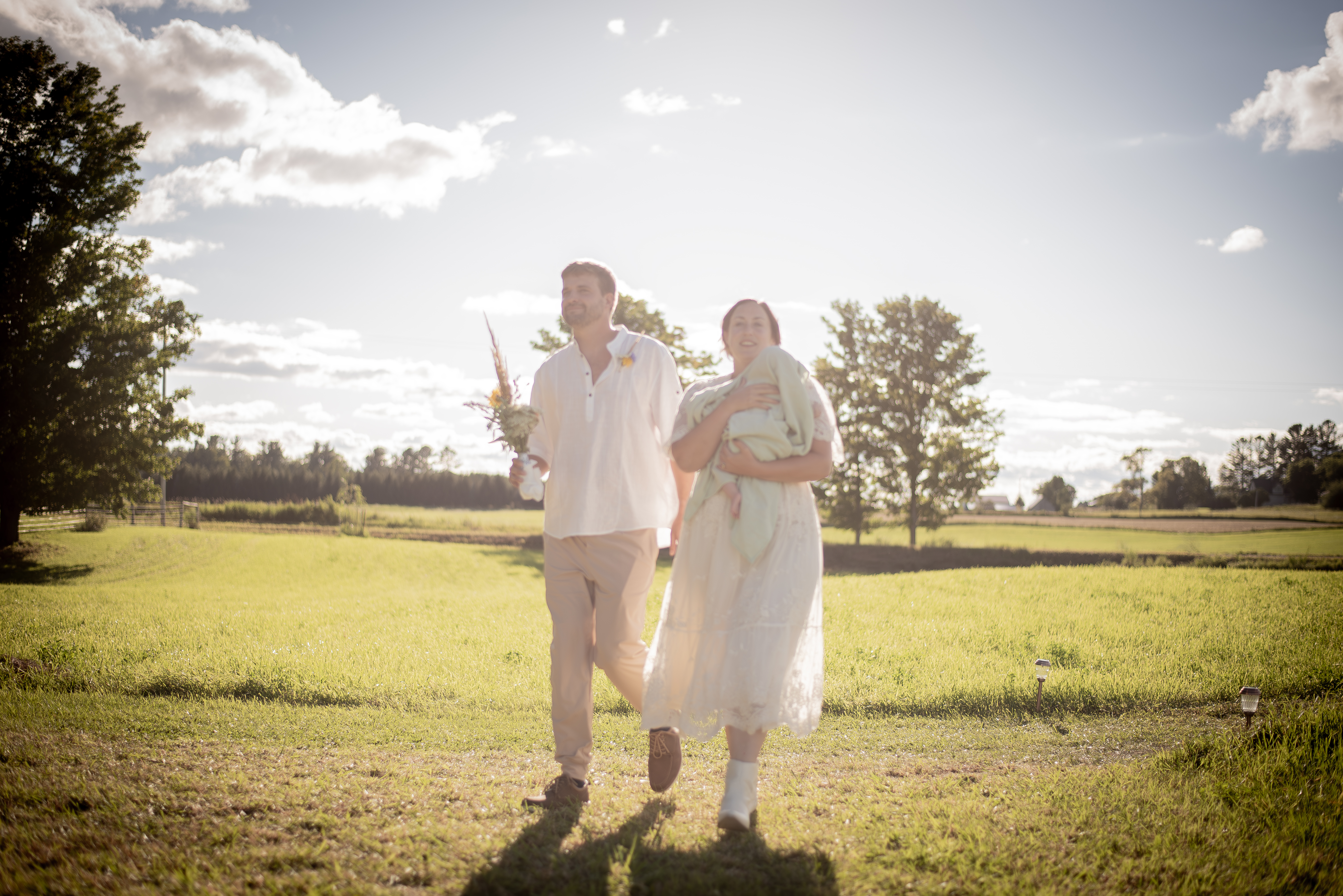 A Young man in light linen and a young woman in a white dress walk toward the camera backed by lighting. The man holds a bouquet and the woman holds a bundle of a baby