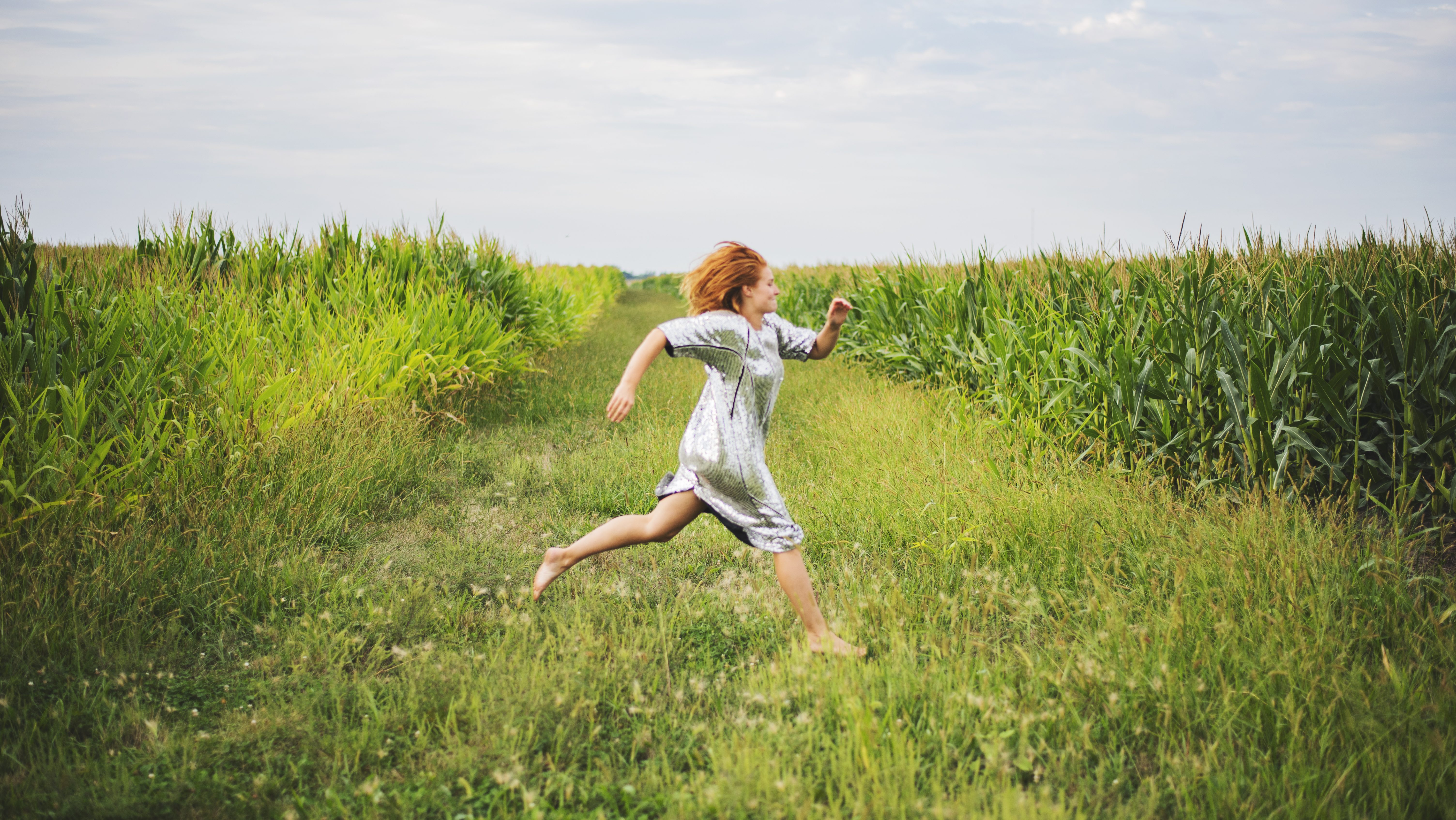 A pale-skinned red headed dancer runs across the frame between rows of tall green corn